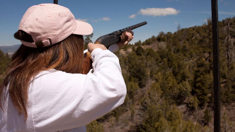 A person wearing a pink cap aims a shotgun at a target in a mountainous outdoor setting.
